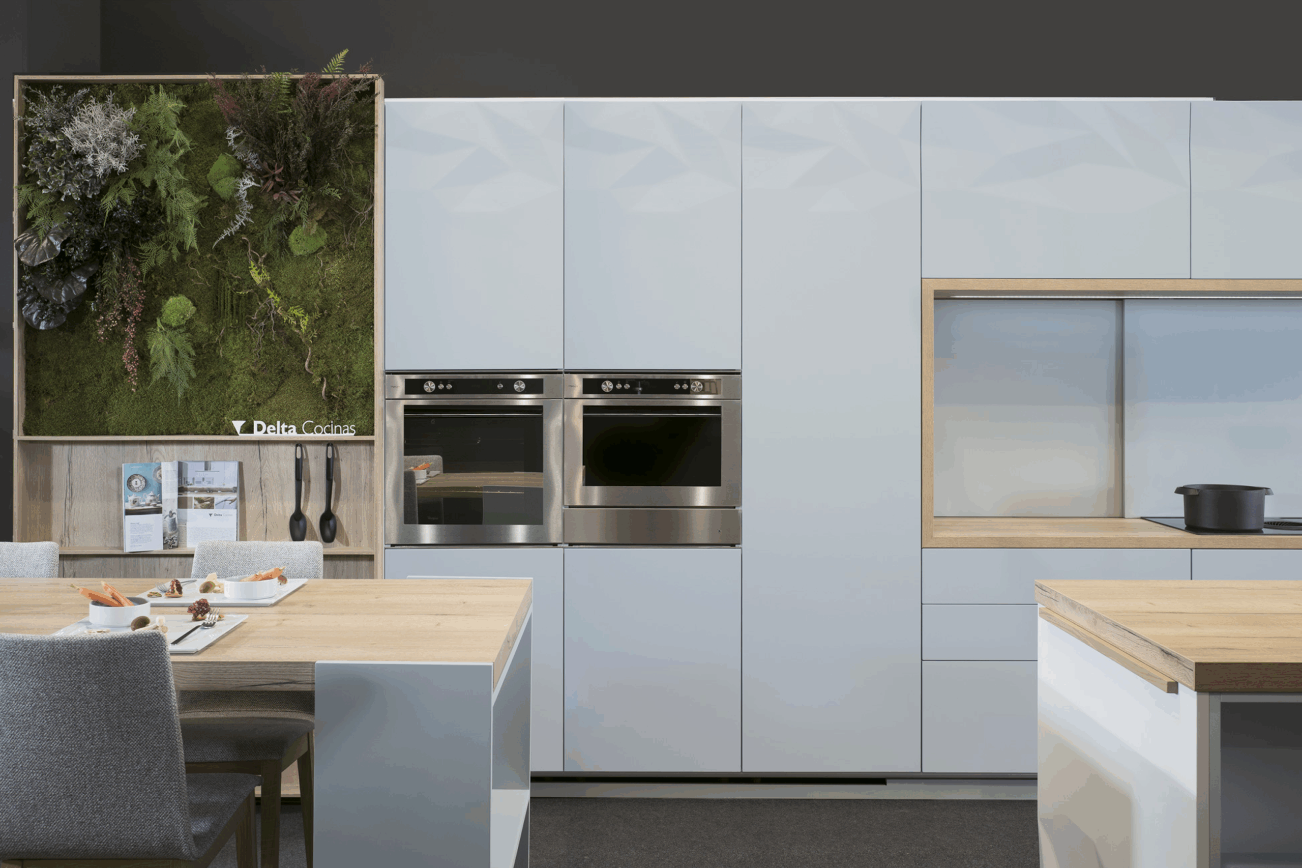 Frentes de armario en acabado blanco mate con textura facetada, integrados con electrodomésticos en acero inoxidable. Panelado continuo con nicho de trabajo enmarcado en madera clara. Diseño pulcro y elegante.