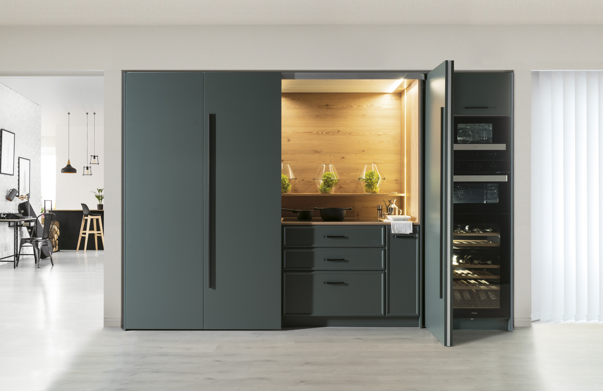 Cocina compacta y funcional con frentes en gris grafito mate y tiradores integrados. El interior, revestido en madera natural, aporta calidez y contraste. La iluminación led bajo estante resalta el fondo y crea una atmósfera acogedora. Las puertas escamoteables permiten ocultarla por completo, ideal para espacios abiertos o multifuncionales.