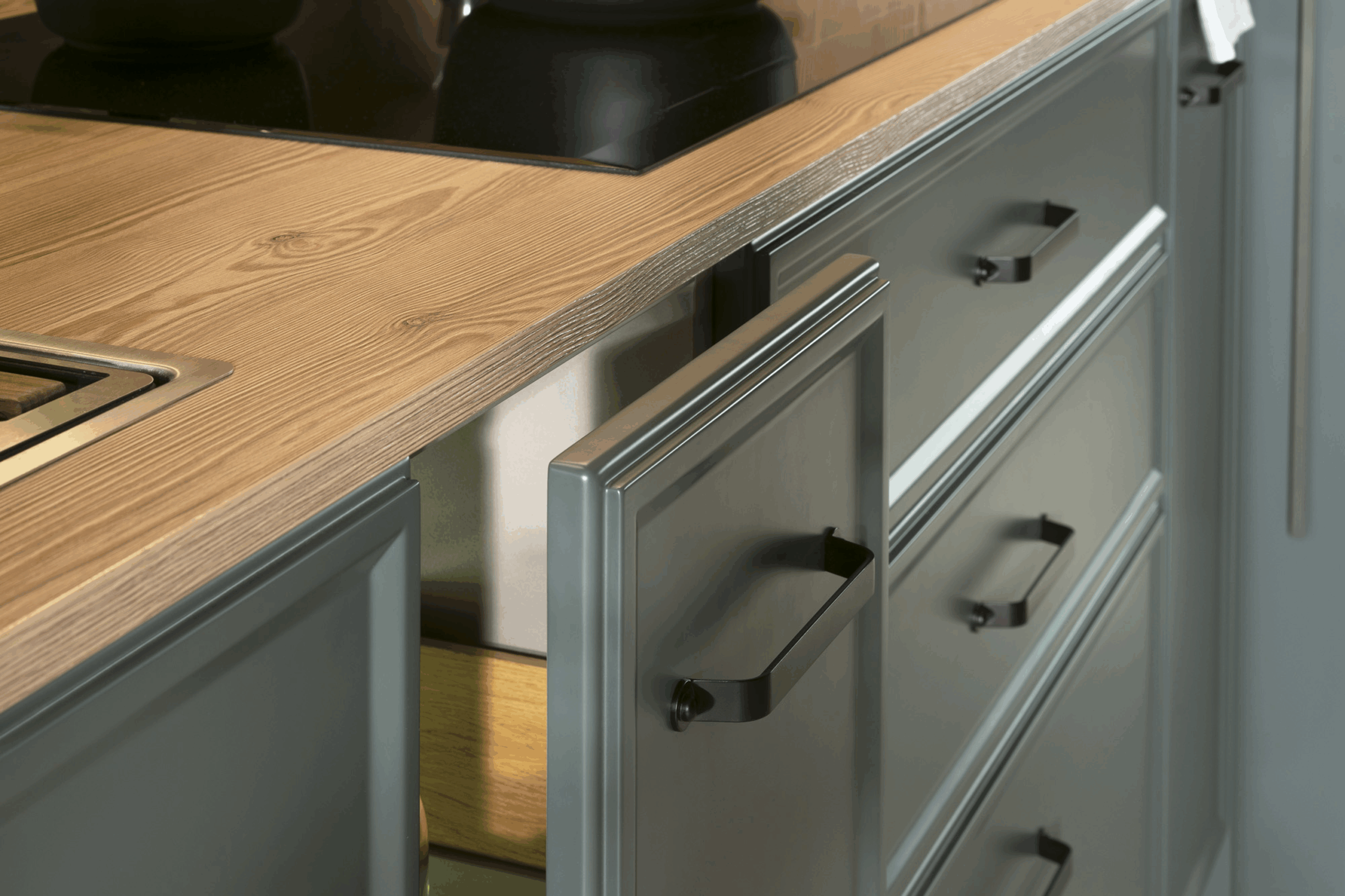 Detalle de cocina con muebles en gris grafito y acabado mate, rematados con tiradores negros metálicos. La encimera de madera natural aporta calidez y contraste al conjunto, mientras que la organización interior revela soluciones funcionales y elegantes para el almacenaje.