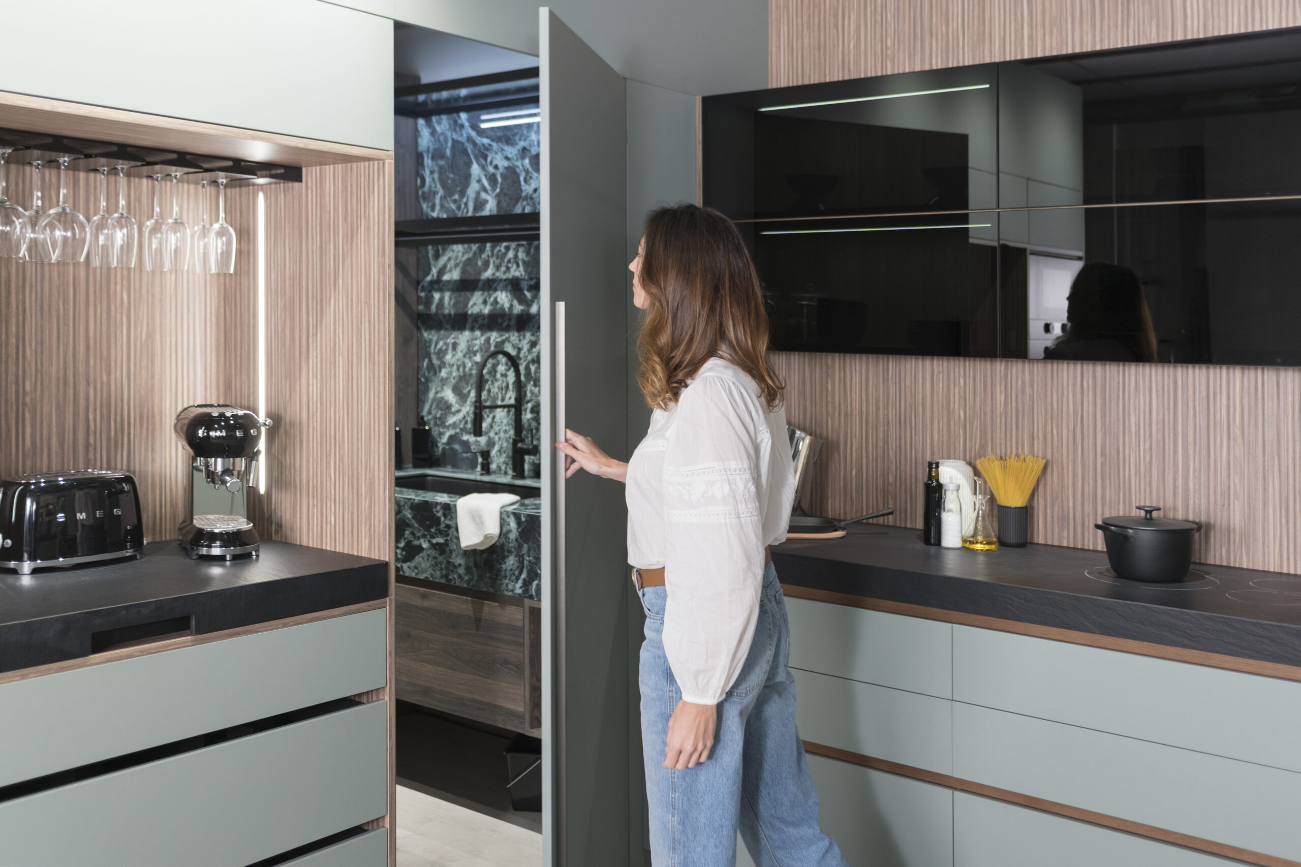 Puerta oculta integrada en el mobiliario de cocina, que conduce a un espacio adicional con acabados en mármol oscuro y grifería negra. La continuidad estética se mantiene gracias a los frentes de mueble en tono verde suave y sin tiradores visibles, acentuando una estética minimalista y funcional.
