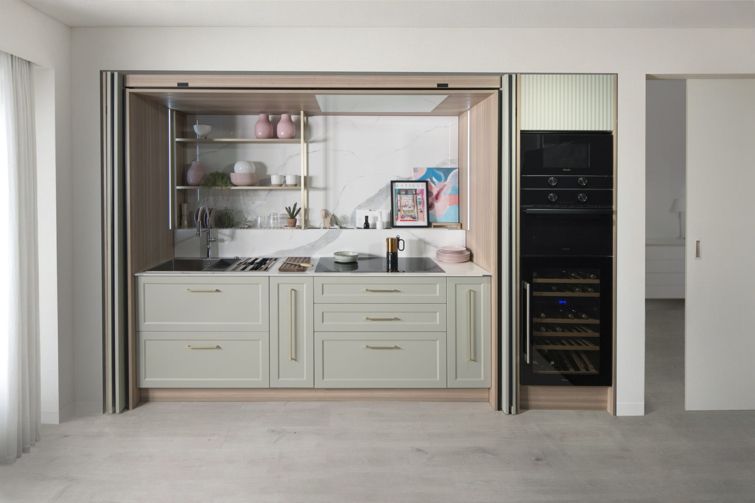 Cocina oculta con muebles en verde salvia y tiradores dorados, que combinan elegancia clásica con funcionalidad moderna. Estantes metálicos dorados, encimera continua y electrodomésticos en columna negra crean un espacio compacto, sofisticado y versátil.