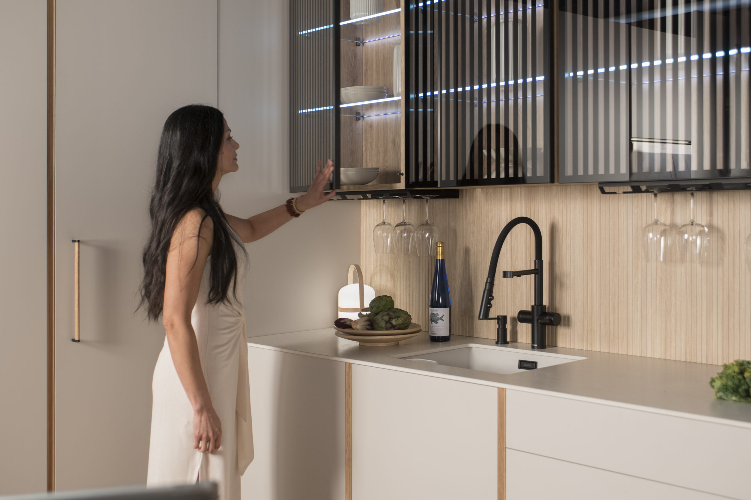 Elegancia minimalista con iluminación tenue. Puertas de vidrio con lamas oscuras revelan vajilla ordenada. Grifería negra y detalles en madera completan un rincón de calma y estilo.